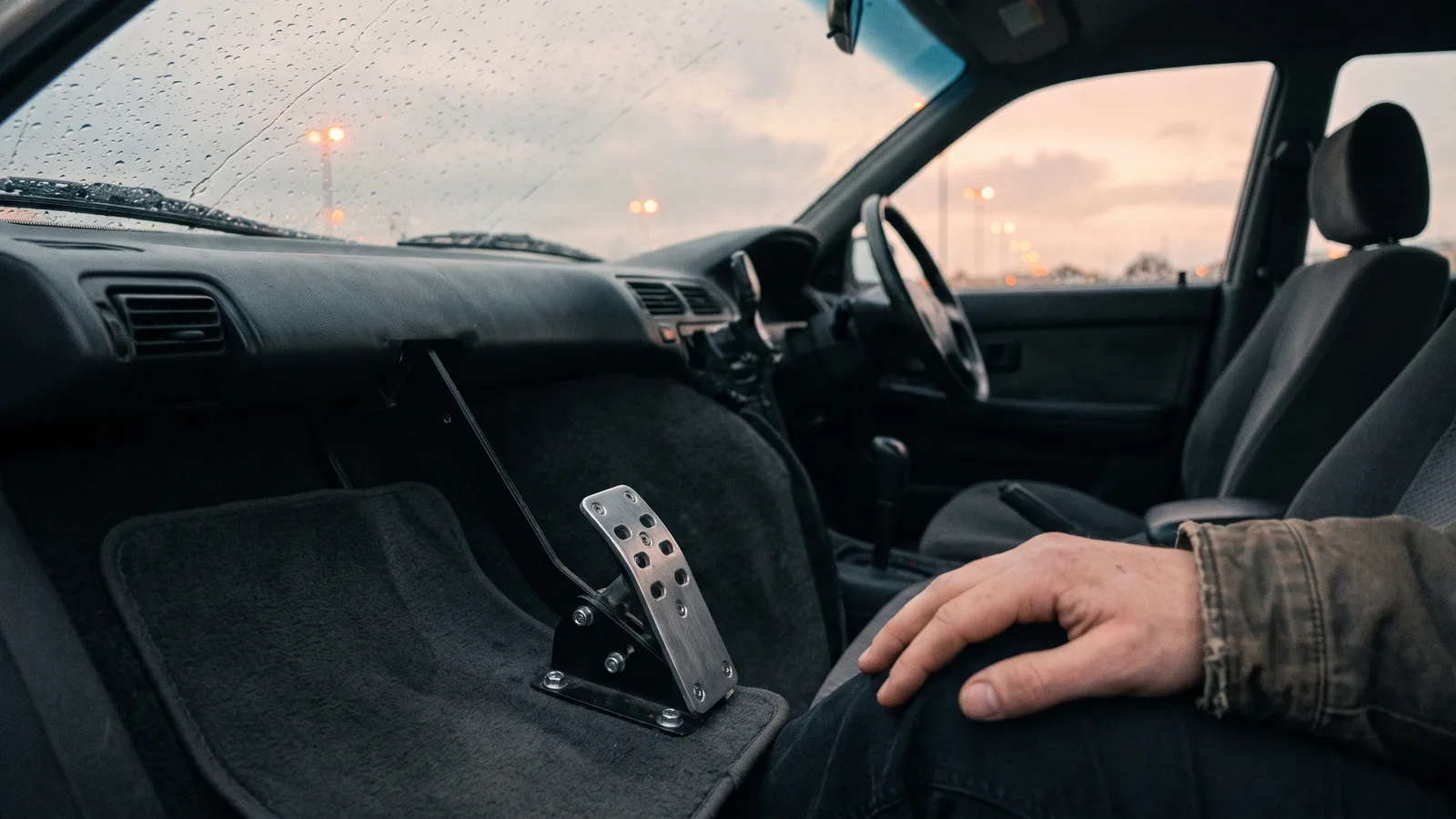 A dual-control training car parked in an empty wet car park at dawn, seen from the passenger side, with a second brake pedal fitted on the instructor's side and a hand resting on the knee above it