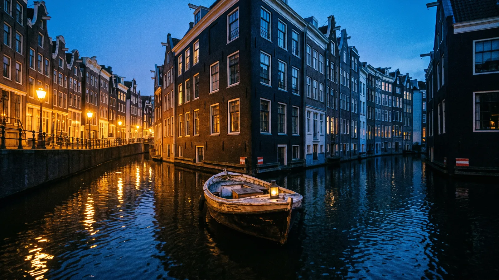 Een bootje bij een splitsing in een Amsterdamse gracht tijdens het blauwe uur: de ene tak is warm verlicht en loopt naar een rij huizen, de andere is koel en loopt naar een andere rij