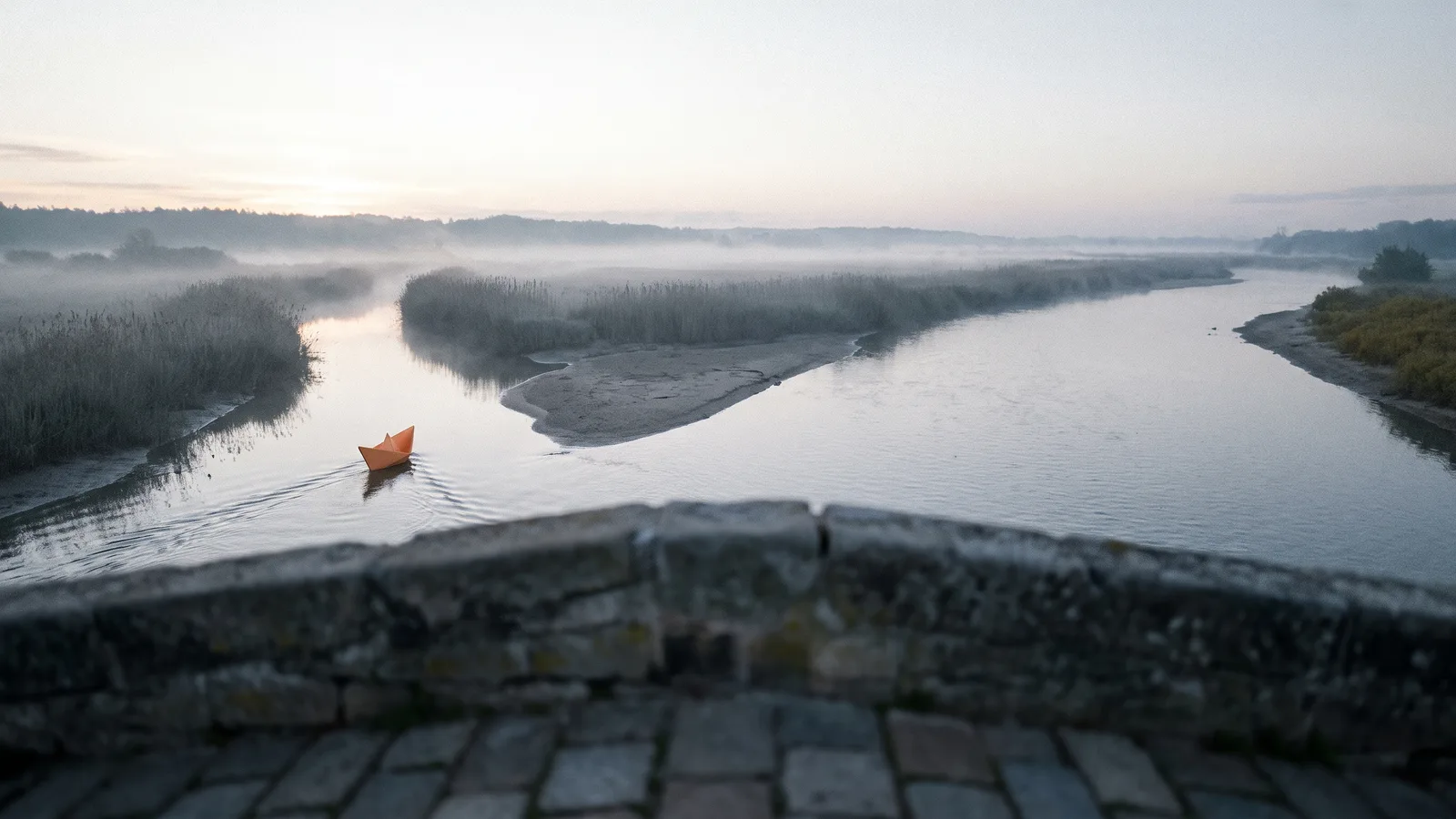 A small orange paper boat drifting into the left channel of a misty river delta at dawn, seen from a low stone bridge