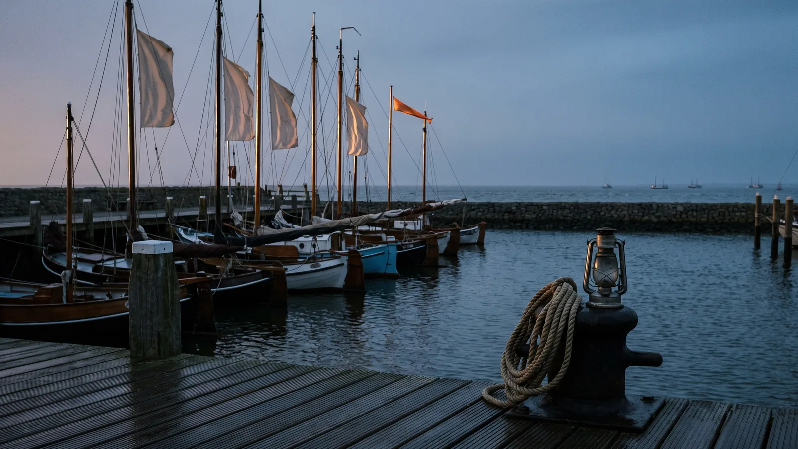 Houten havensteiger in de schemering met een rij afgemeerde bootjes, waarvan één met een opvallende oranje wimpel tussen gewone witte vlaggen