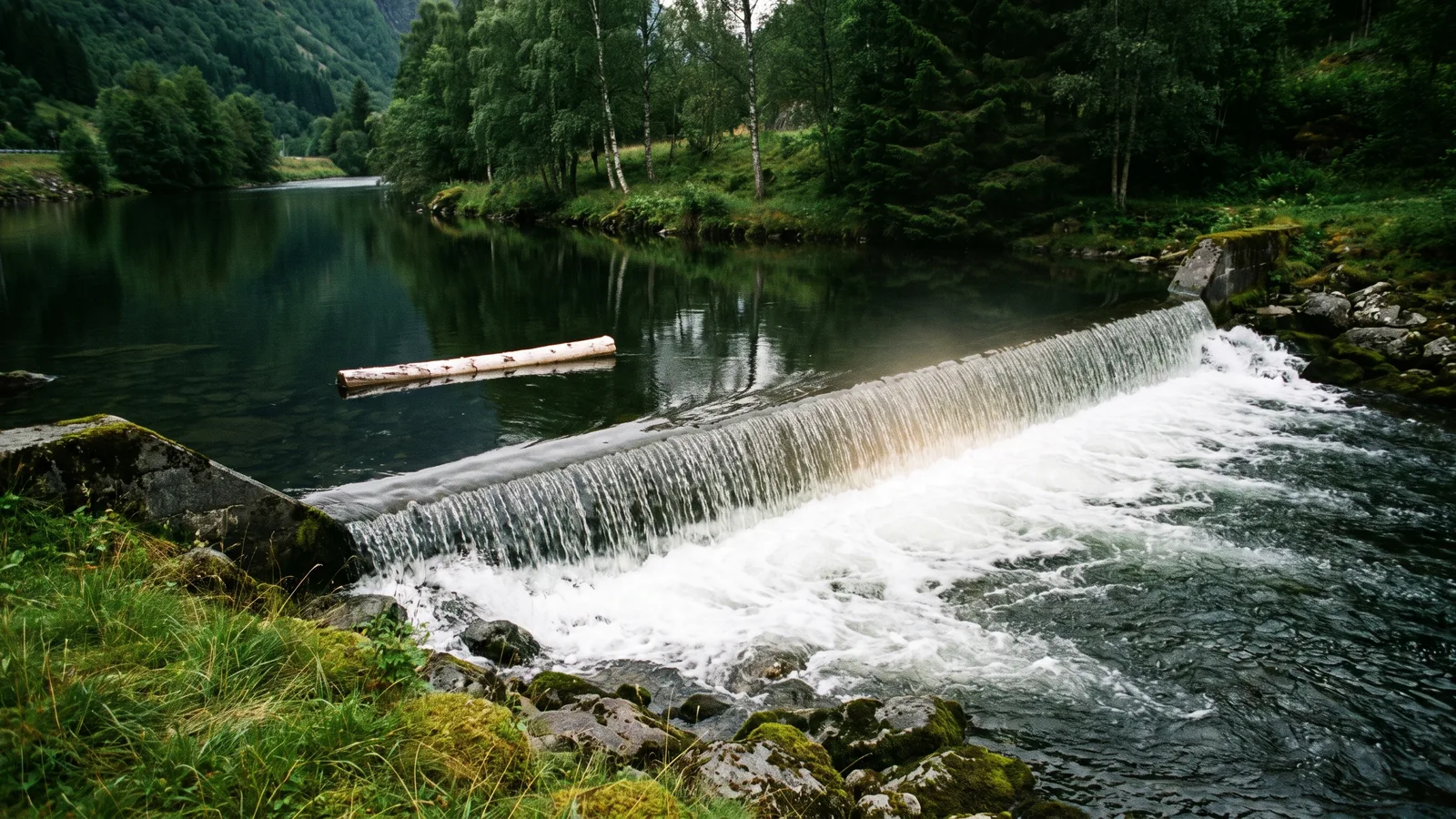 Een lage betonnen stuw in een rivier in een groen Noors dal, met kalm donker water erboven, wit schuimend water eronder en één boomstam die naar de rand drijft